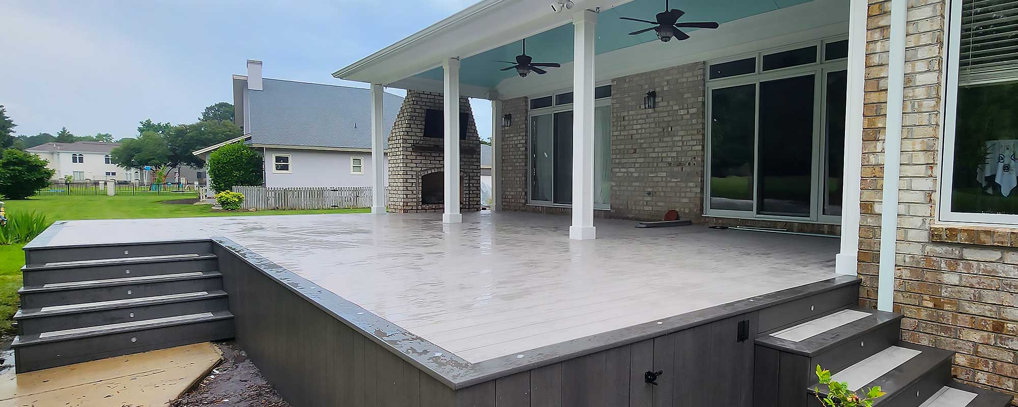 A spacious backyard features a high-quality custom deck with composite decking, covered by a roof with ceiling fans. The deck is connected to a brick…| First In Decks and Fencing | Virginia Beach Deck Builders