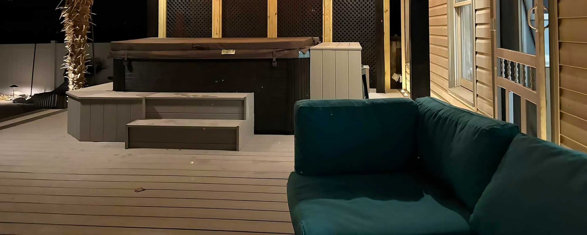 A cozy outdoor living space features a hot tub on a custom deck with steps, surrounded by a privacy screen. A dark teal sofa sits in the foreground, highlighting professional craftsmanship and the use of durable materials for home improvement.| First In Decks and Fencing | Virginia Beach Deck Builders