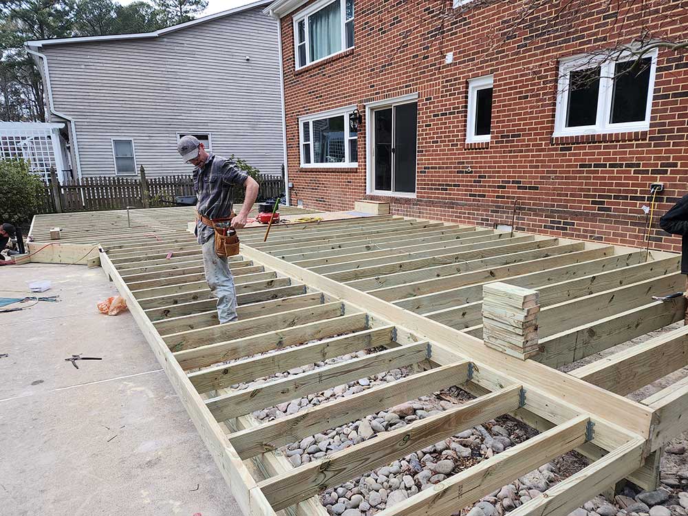 A worker installs a custom deck using pressure treated lumber in a backyard, showcasing the beginning stages of a backyard transformation. The deck frame is…| First In Decks and Fencing | Virginia Beach Deck Builders