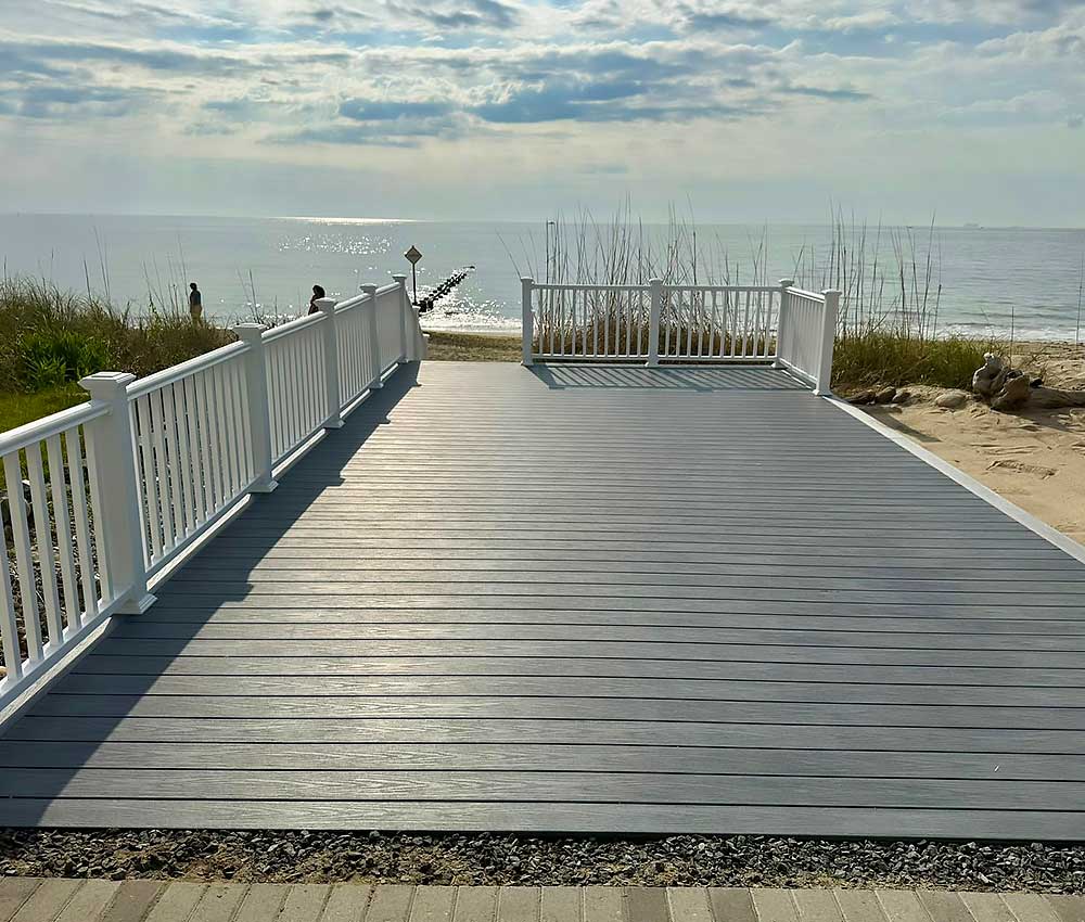 A scenic view of a gray composite decking overlooking a beach, featuring white railings and surrounded by sand dunes, exemplifying high-quality, low-maintenance outdoor living with durable materials for a backyard transformation.| First In Decks and Fencing | Virginia Beach Deck Builders