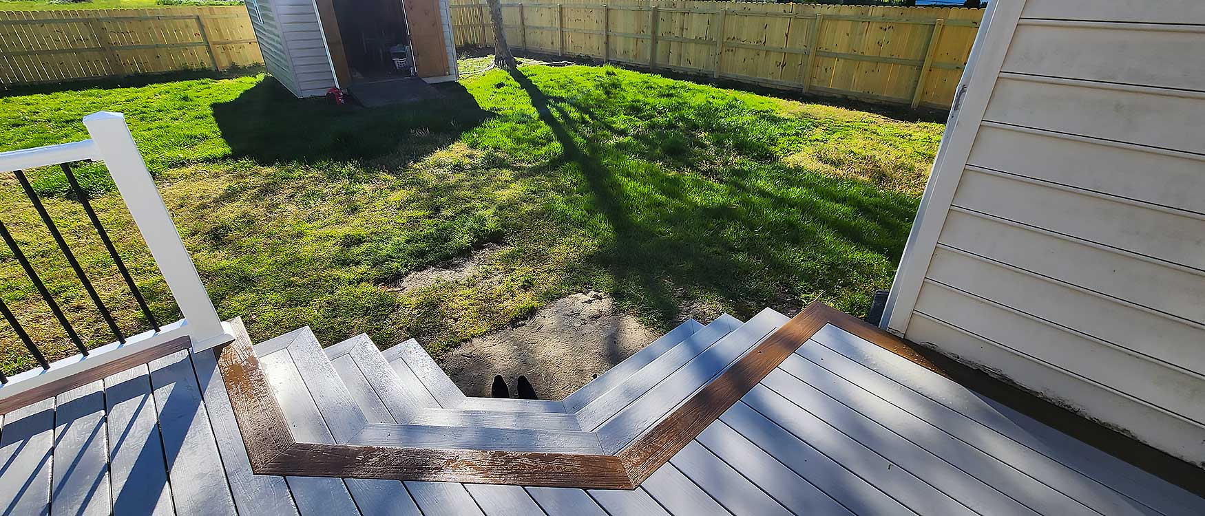 View from a composite deck with gray steps leading to a green backyard, enclosed by a wooden fence, highlighting a shed in the corner, ideal for outdoor living and showcasing durable materials for home improvement.| First In Decks and Fencing | Virginia Beach Deck Builders