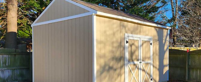 A beige shed with white trim, located in a backyard, features a small wooden ramp leading to double doors. Surrounded by trees and a wooden…| First In Decks and Fencing | Virginia Beach Deck Builders