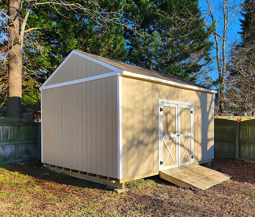 A beige shed with white trim, located in a backyard, features a small wooden ramp leading to double doors. Surrounded by trees and a wooden…| First In Decks and Fencing | Virginia Beach Deck Builders
