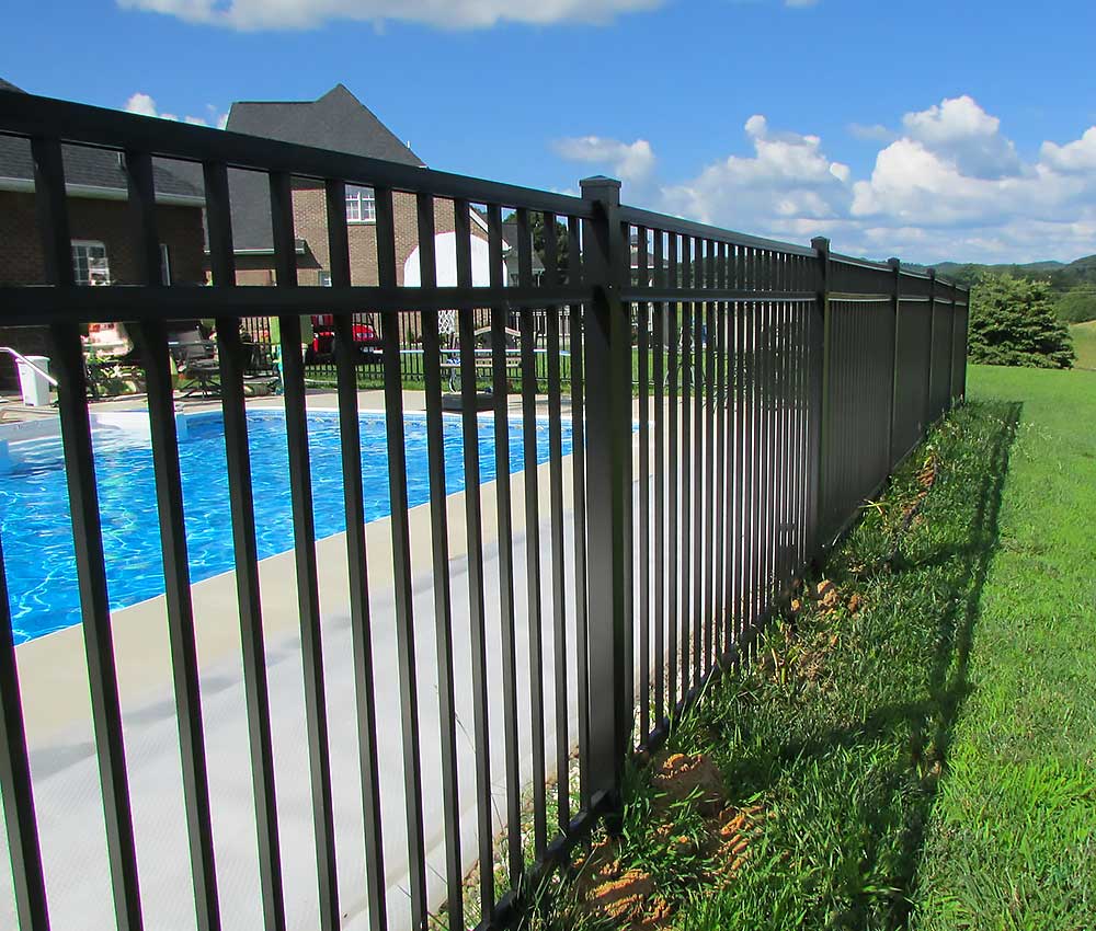 A black metal fence encloses a swimming pool next to a brick house under a blue sky. The scene highlights professional craftsmanship and durable materials, ideal for outdoor living and fence installation projects in Chesapeake.| First In Decks and Fencing | Virginia Beach Deck Builders