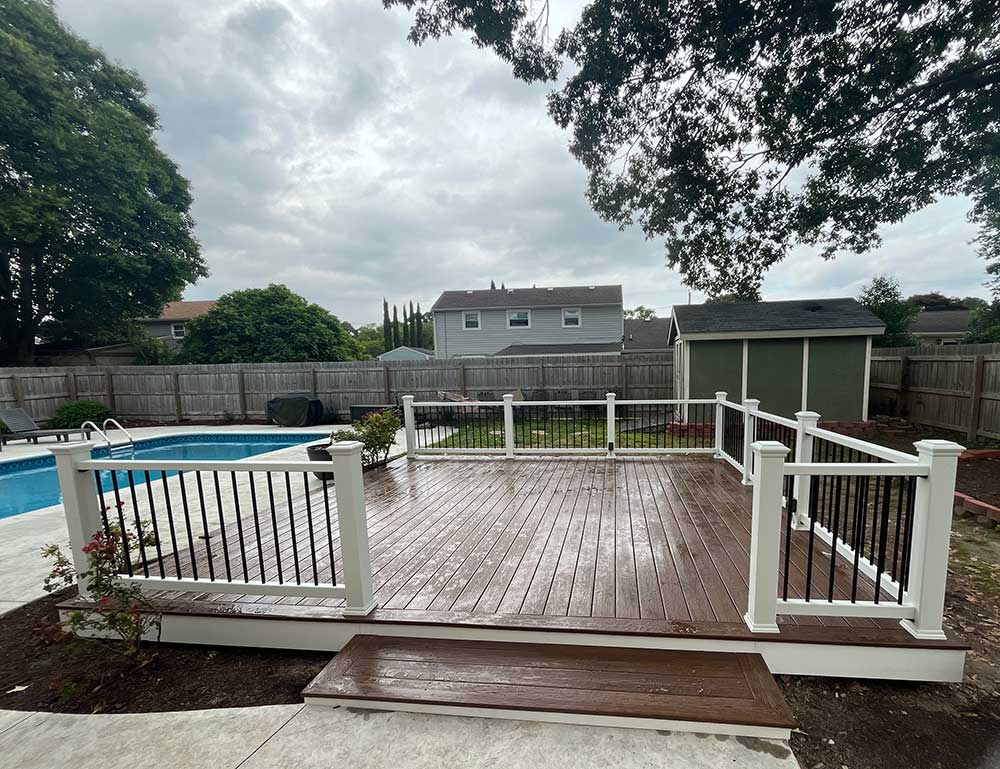 A spacious backyard features a newly installed custom deck made of durable materials, overlooking a swimming pool and surrounded by a wooden fence. A shed is visible in the background, exemplifying professional craftsmanship in outdoor living.| First In Decks and Fencing | Virginia Beach Deck Builders