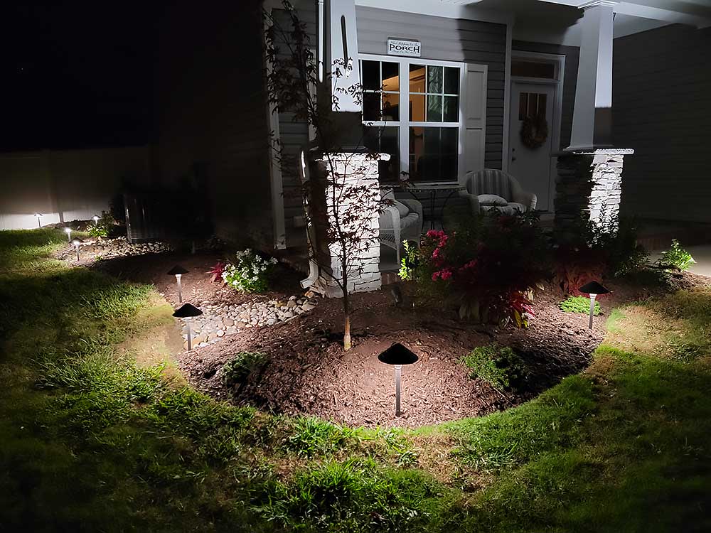A beautifully lit porch at night features cozy chairs and a neatly landscaped garden, highlighting outdoor living. The scene emphasizes professional craftsmanship and durable materials,…| First In Decks and Fencing | Virginia Beach Deck Builders