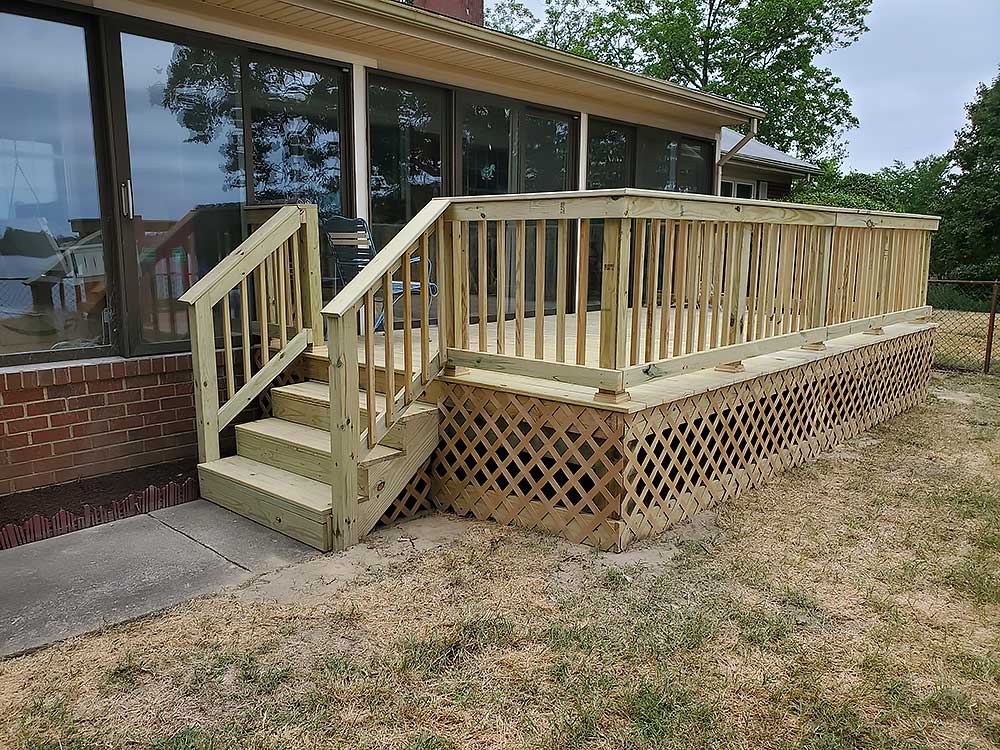 A newly constructed wooden deck with railing and lattice skirting extends from a brick house, showcasing professional craftsmanship and durable materials, ideal for enhancing outdoor living in Chesapeake with custom decks.| First In Decks and Fencing | Virginia Beach Deck Builders