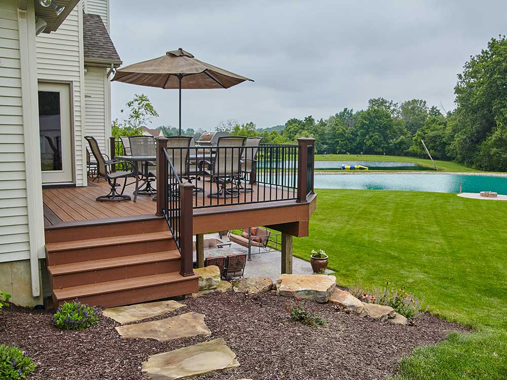 Chesapeake-VA-image-4-mobile A spacious custom deck made of high-quality composite decking overlooks a lush backyard with a pond, featuring outdoor furniture and an umbrella, showcasing professional craftsmanship and promoting outdoor living.| First In Decks and Fencing | Virginia Beach Deck Builders