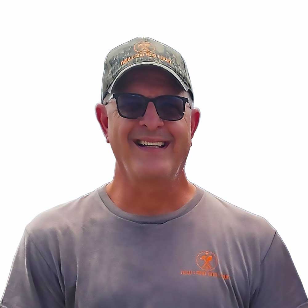 A smiling man in sunglasses and a cap stands outdoors, wearing a gray shirt with an orange logo. He represents professional craftsmanship in custom decks…| First In Decks and Fencing | Virginia Beach Deck Builders