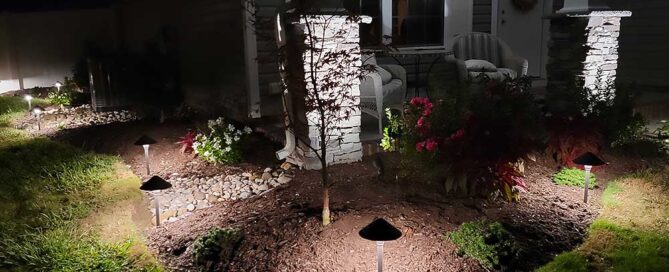 A well-lit front porch at night, featuring a landscaped garden with small trees and flowers. The scene highlights outdoor living enhancements, perfect for backyard transformation and home improvement projects in Chesapeake.| First In Decks and Fencing | Virginia Beach Deck Builders