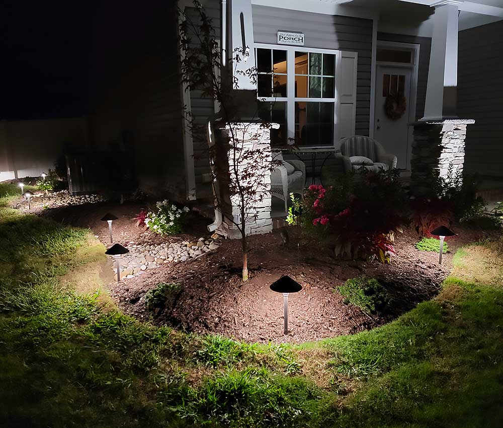 A well-lit front porch at night, featuring a landscaped garden with small trees and flowers. The scene highlights outdoor living enhancements, perfect for backyard transformation and home improvement projects in Chesapeake.| First In Decks and Fencing | Virginia Beach Deck Builders